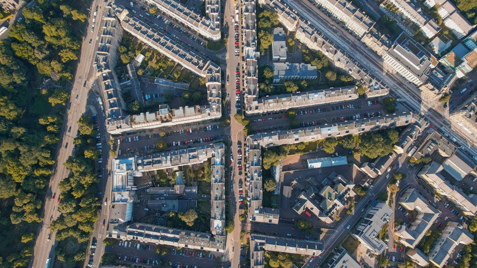 An aerial view of a densely populated urban area showing rows of terraced residential buildings with grey rooftops, intersected by a central road with parked cars on either side. In the foreground, a 